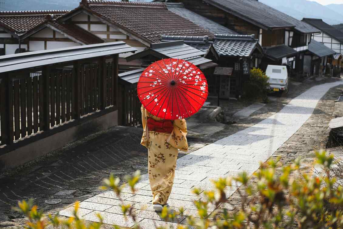 Person in traditioneller, asiatischer Kleidung läuft mit rotem Sonnenschirm eine Gasse entlang.