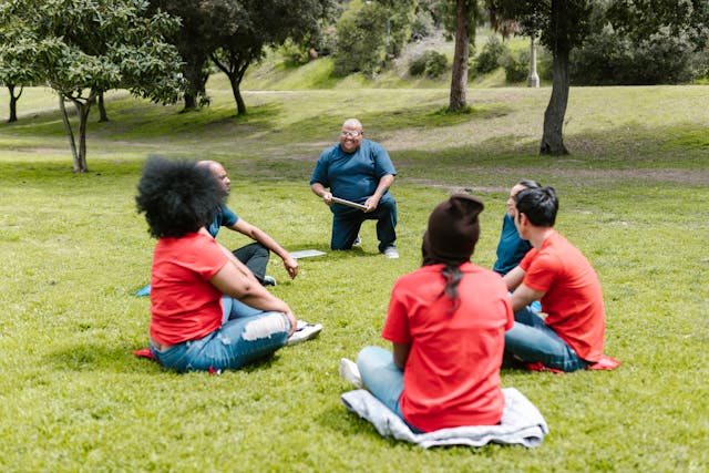 Eine Gruppe von Mitarbeitern sitz im Park und hört dem Teamleader aufmerksam zu.